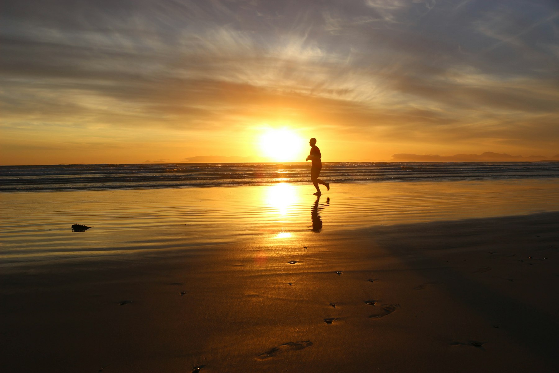 A runner silhouetted on the beach at sunset, reflected in the wet sand